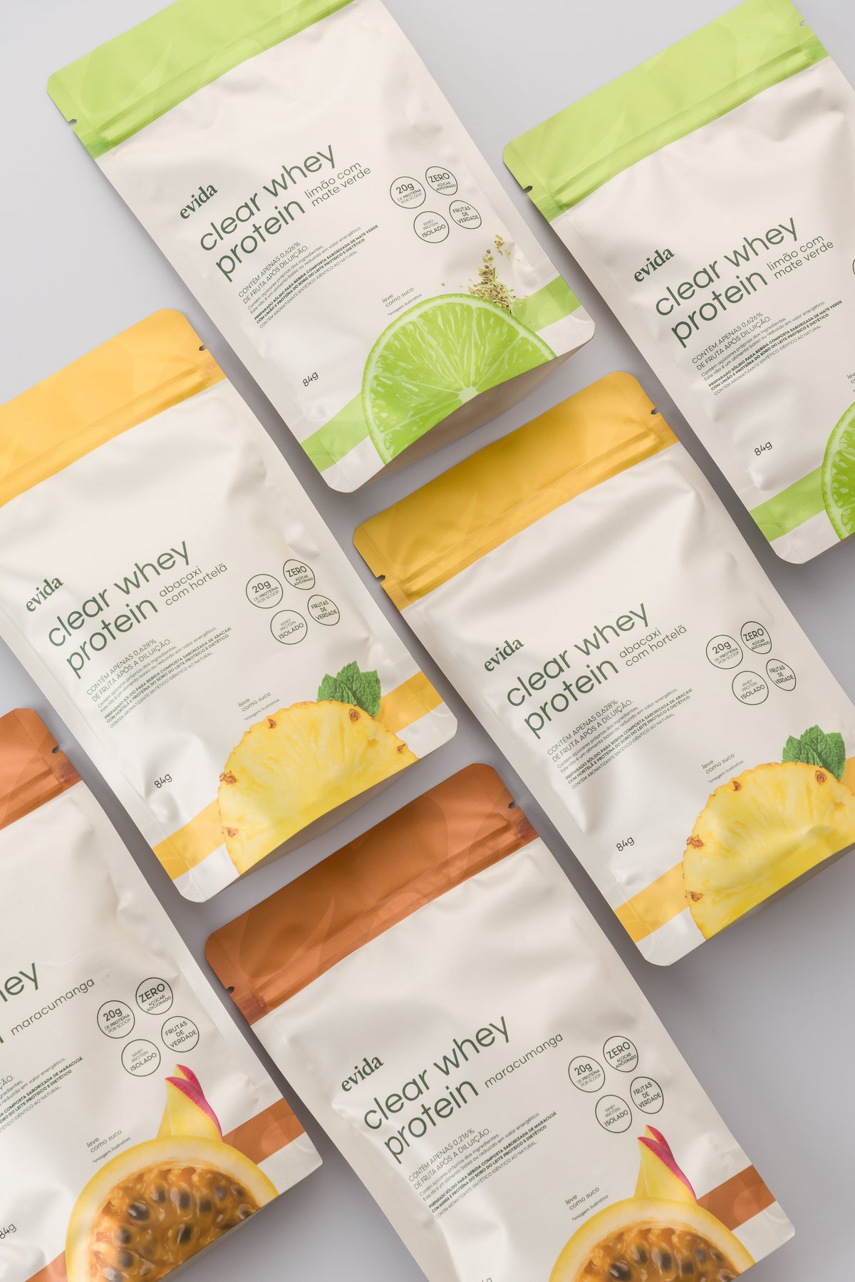 Sachês de clear whey protein da Evida espalhados sobre fundo claro, nos sabores abacaxi com hortelã, limão e maracujá com manga
