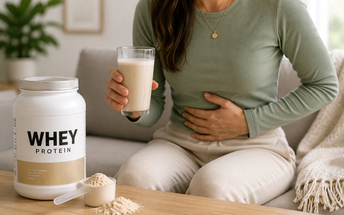 Mulher sentada no sofá segura um copo de whey protein com a outra mão sobre a barriga, com pote e dosador de whey na mesa à frente