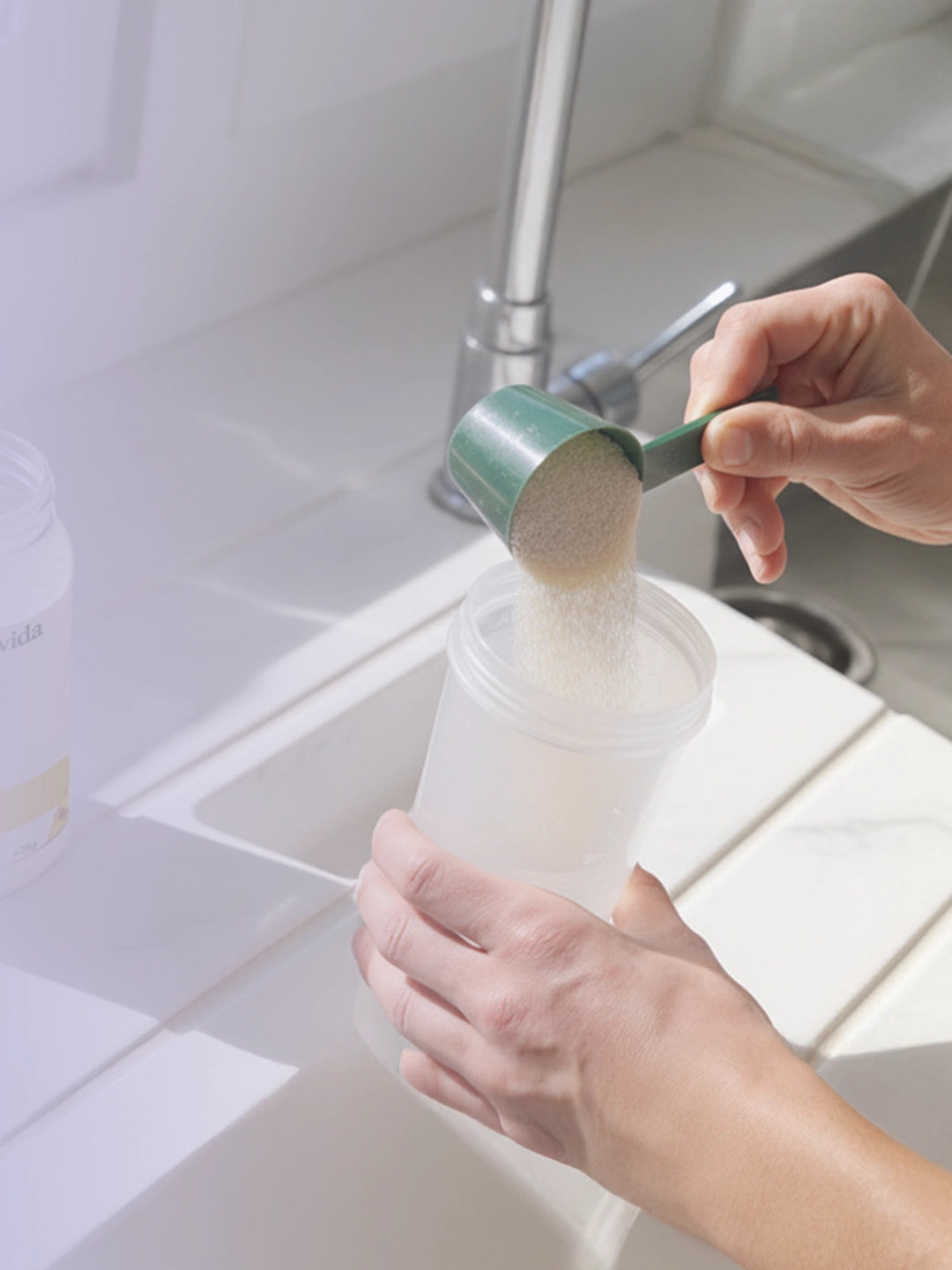 Person pouring a scoop of white powder into a clear plastic shaker cup near a kitchen sink.