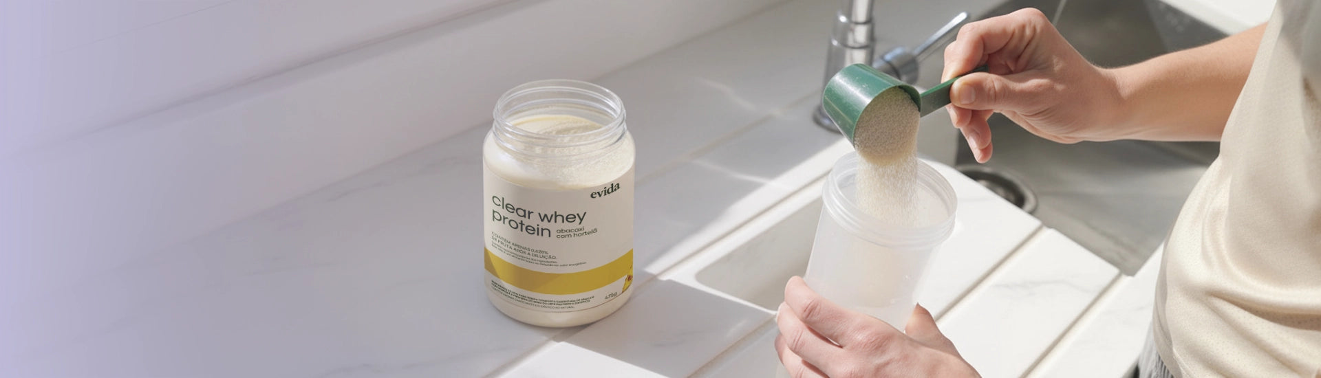 Person scooping powder from a container into a clear shaker bottle on a kitchen counter with sunlight.