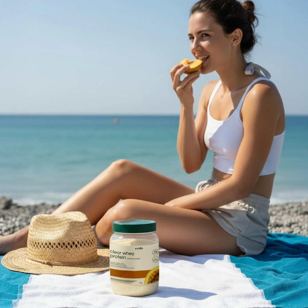 Young woman in white top and shorts sitting on a beach towel by the sea, eating a snack with a straw hat nearby